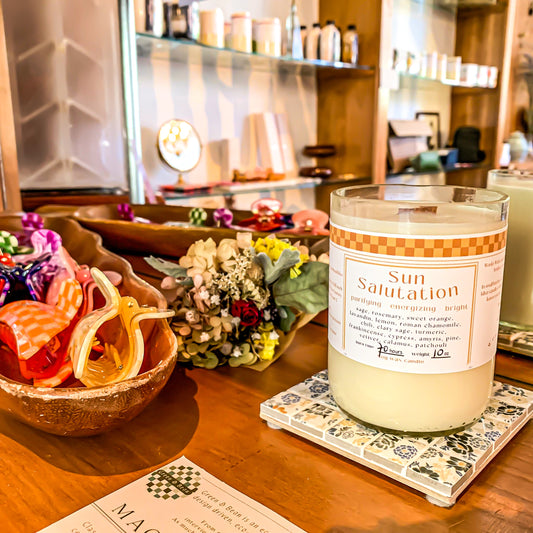 Sun Salutation candle on a shelf display next to hair claws. Candle label shows the scent is "purifying energizing and bright" with a list of essential oil ingredients listed. Includes sage, rosemary, lemongrass.
