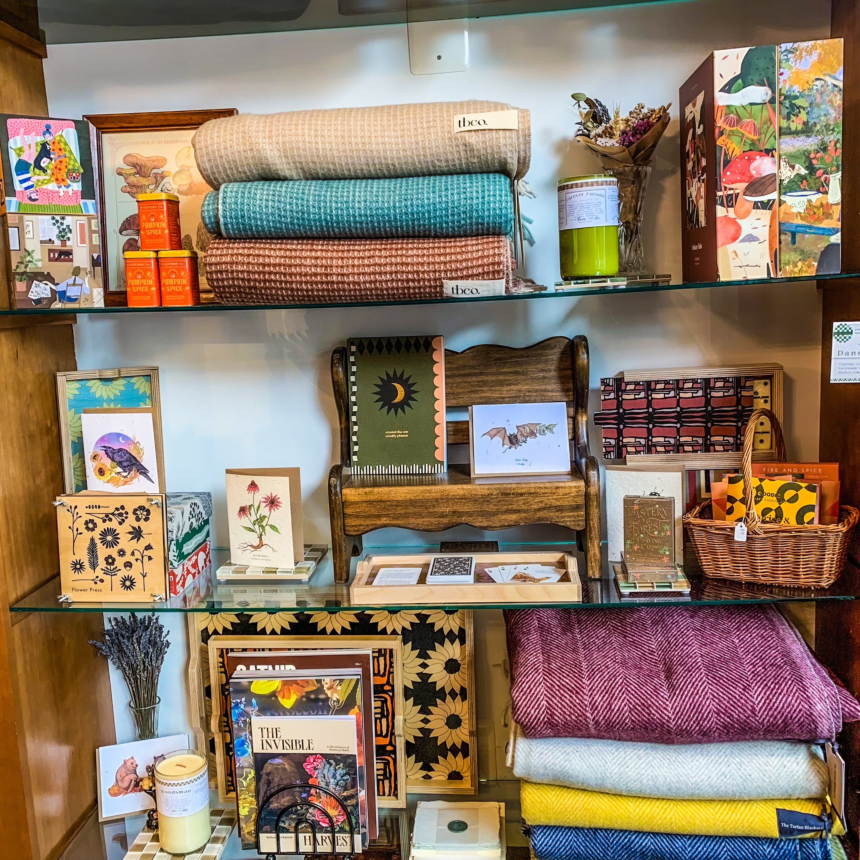Green & Bean gift shop shelf display for fall. Brom bottom to top there are recycled wool blankets, a stack of magazines, xoy candles, dried lavender, a wicker basket of vegan chocolates, a mini wooden display bench with a bat greeting card, fall puzzles and a flower press. 