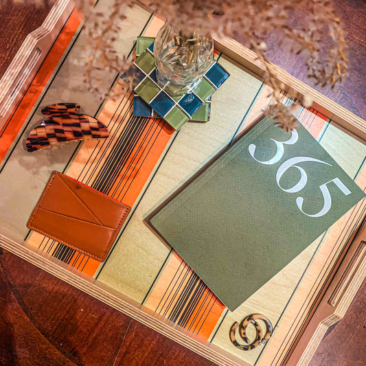 Coffee table tray display featuring 365 planner in moss, a vegan brown wallet, blonde tortoise hoops and hair claw, and a blue/green checkered glass tile with a glass vase and dried flowers. Tray is green with peach and orange vertical sections and thin black stripes.