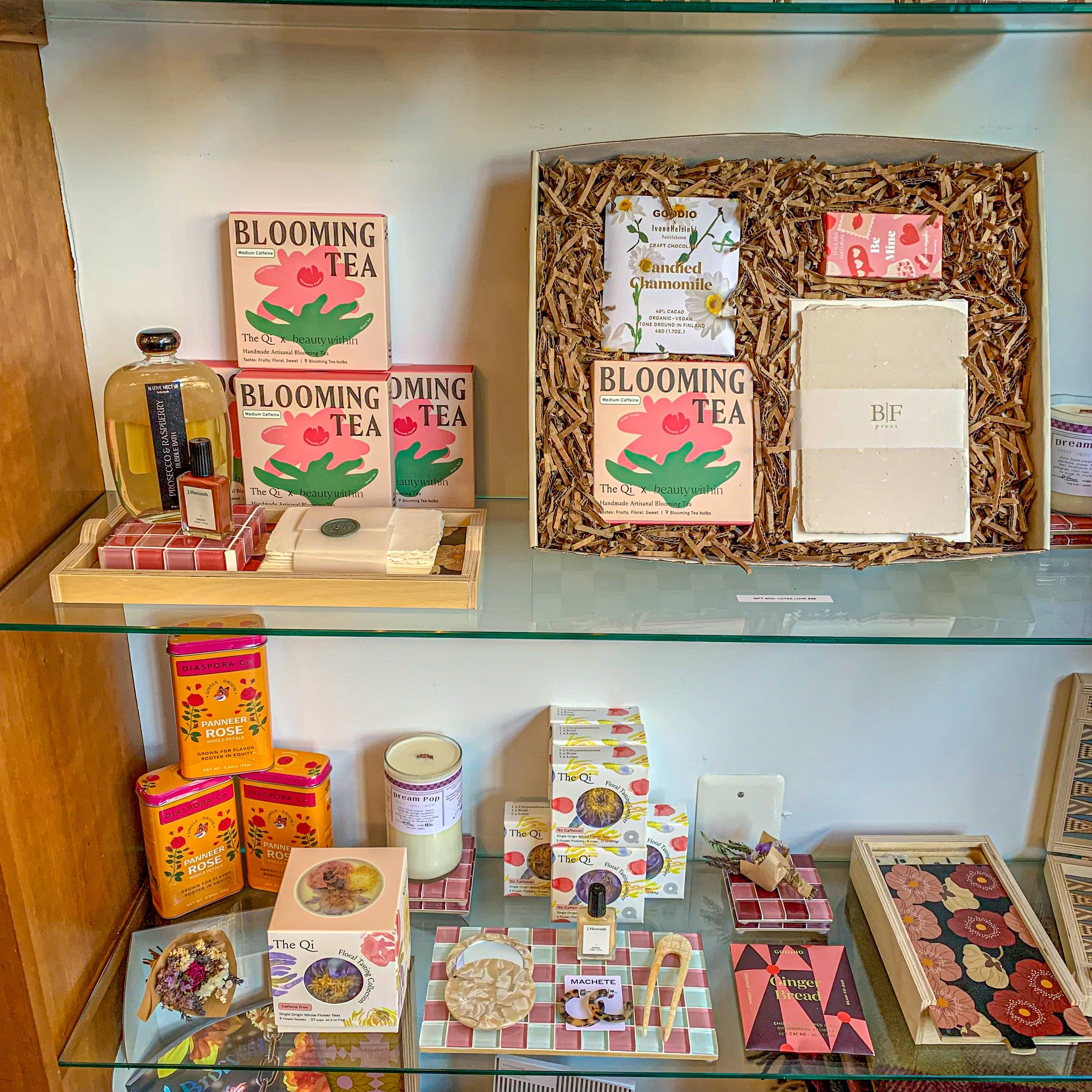 Green & Bean boutique valentines day shelf display with pink motif. Products include blooming tea set, pink & white checkered tile tray, floral domino set, Diaspora Co. Panneer roses, nail polish, and a curated gift box.