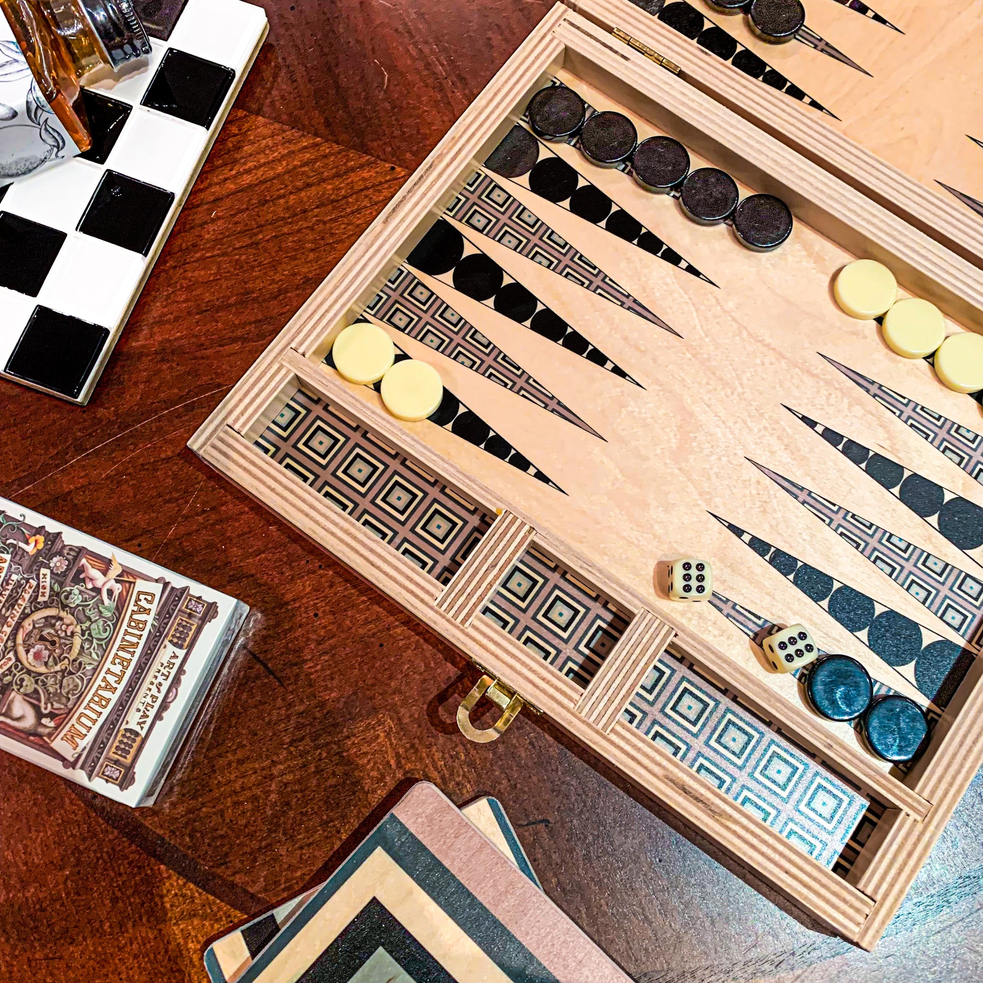 Close up of Squaresville Grey backgammon set with the traditional game set up. Points are alternating black circles and grey squares. Matching coasters, a deck of playing cards and a black and white trivet line the picture for situational context of a game night