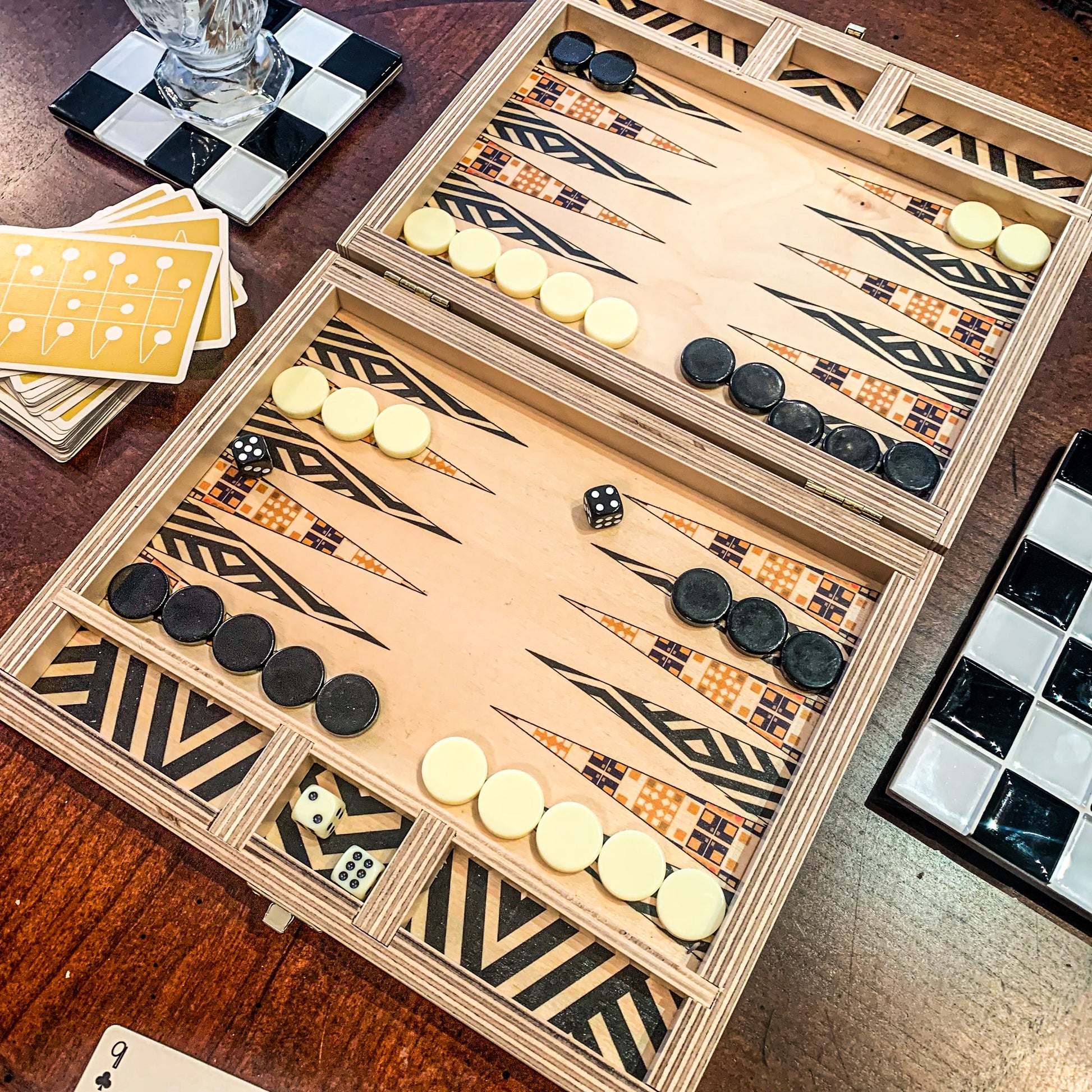 Backgammon game set up on a wooden travel set by Solfum Studio in the Alexander Olive pattern. Contrasting black & white stripes and a mid-century inspired patter make up the points and the black dice are rolled to 4 and 5