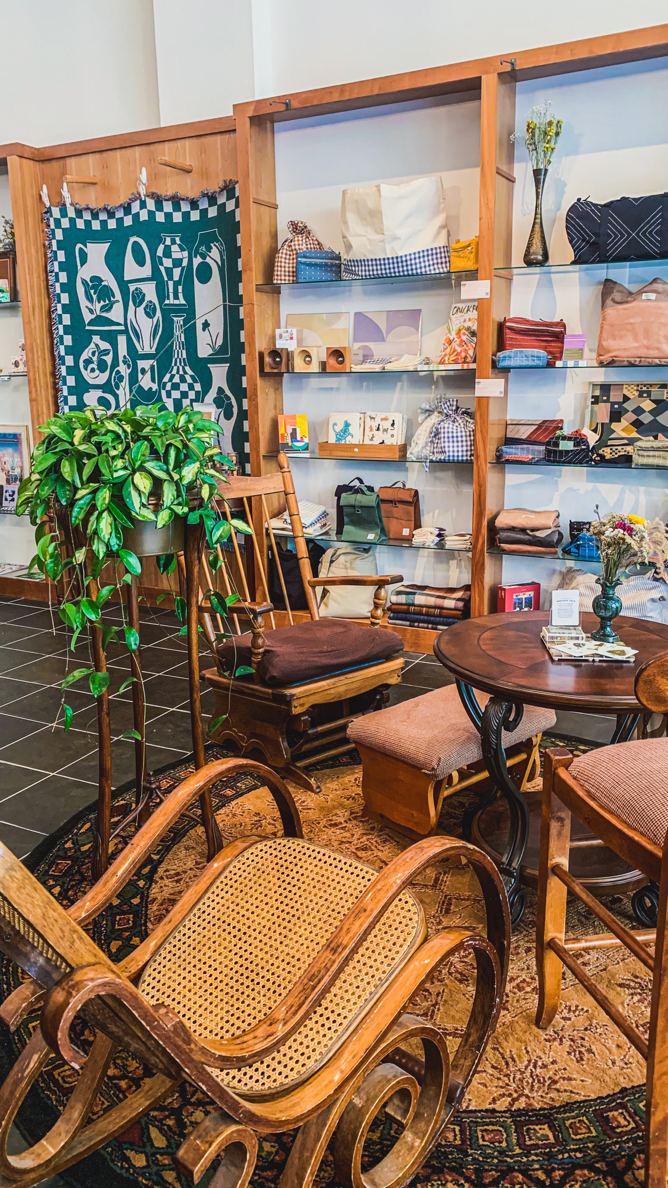 Green & Bean gift shop interior focused on the Bentwood rocking chair in the store seating area. In the background a green tapestry hangs behind a large hoya in a plant stand and shelves of colorful product can be seen