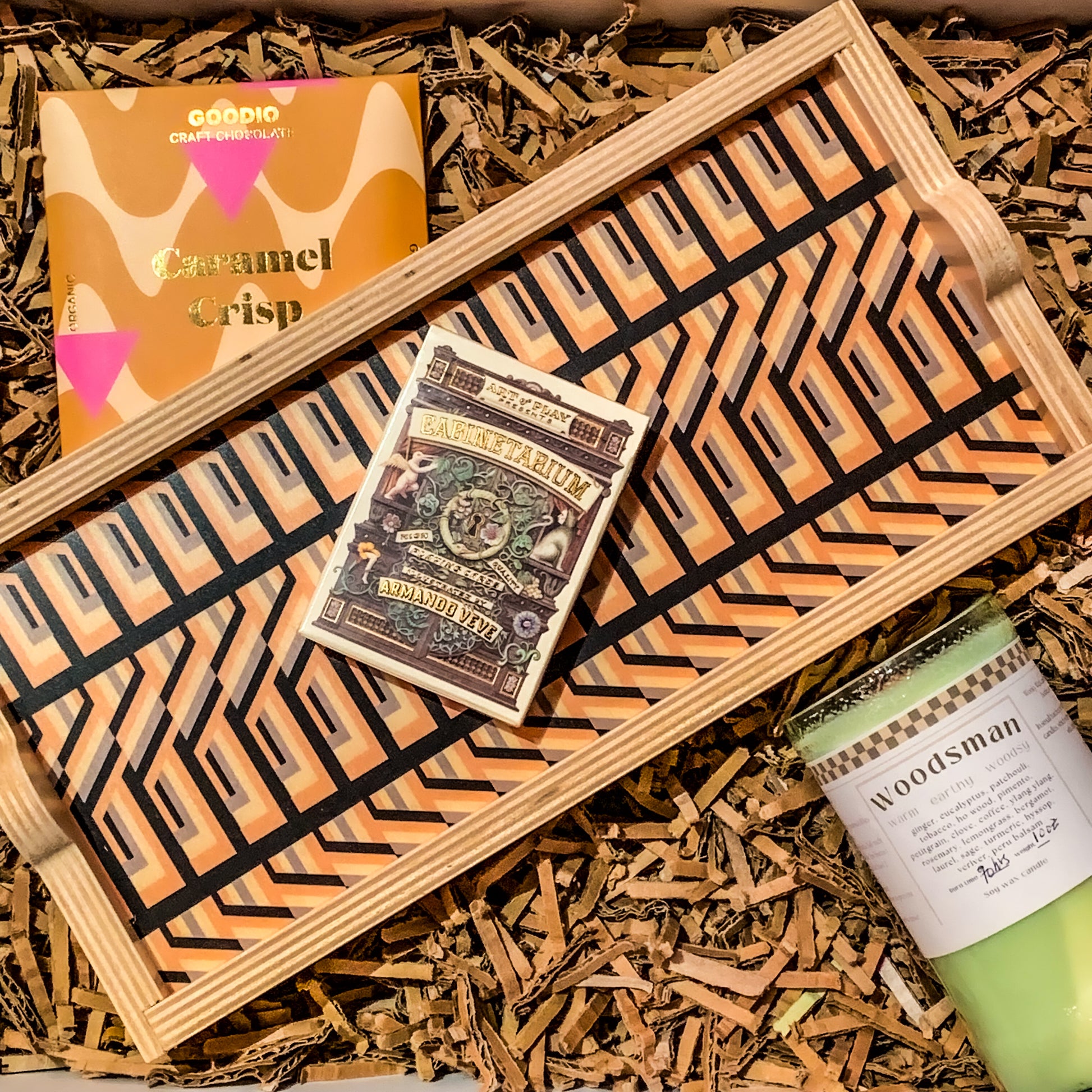 Variation B of Ali's Housewarming gift box: a graphic striped pattern tray with a "cabinetarium" deck of playing cards, vegan caramel chocolate, and a woodsy soy wax candle
