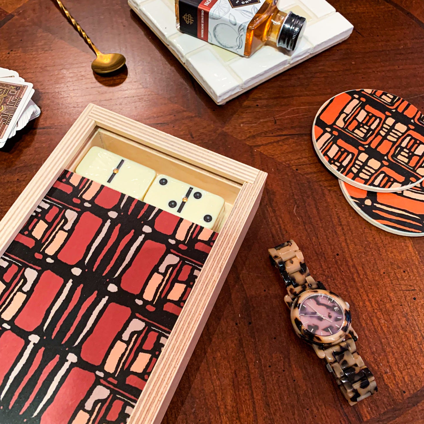 luxury domino set in rue maroon with matching pair of circular coasters, a blonde tortoise watch, a golden bar spoon add to the background of the photo set up, but the Domino is the focal point