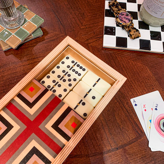Games night set up with a focus on a wooden domino set. The box has a top that slides open to reveal the dominoes beneath and has a red T and smaller layers of squares. There is also 3 playing cards in the right corner, and a checkered tray with a watch on top.