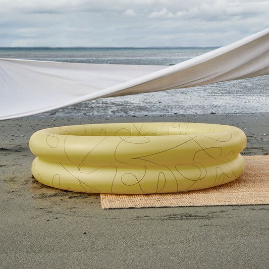 Mylle Inflateable Pool in Lines is a solid yellow color with thin black abstract lines. Displayed on a beach with a sheet of fabric stretching the top of the photo artistically