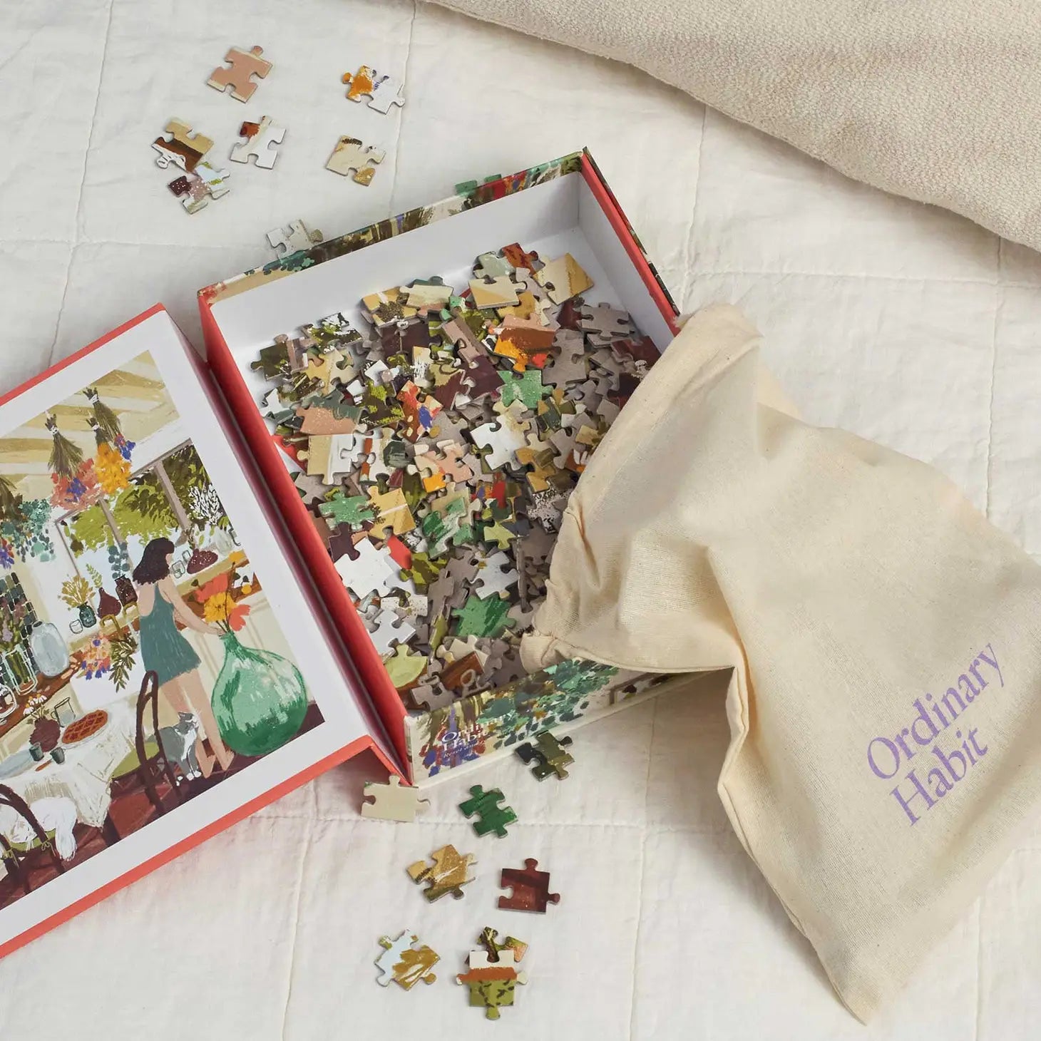 Open box display of Ordinary Habit's "Home Flowering" 500 piece puzzle. Shows inside the puzzle box with the puzzle picture on the left side depicting a girl arranging dried flowers in her kitchen. There is a drawstring bag to contain the pieces which are spilling out of the bag into the box.