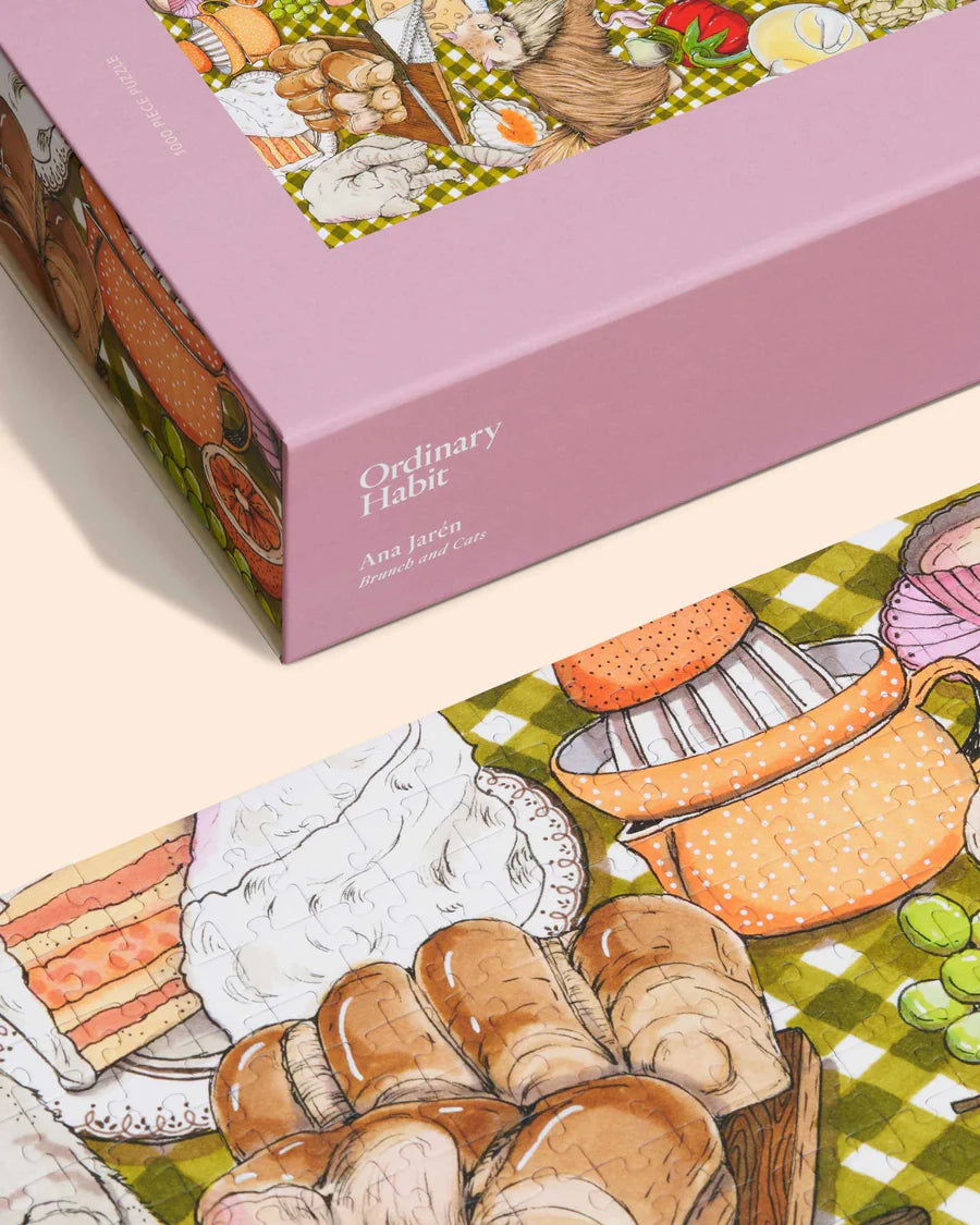 Corner view of Ordinary Habit's "Brunch and Cats" Puzzle. Tjos section shows a sliced cake, hot cross buns, and a orange on a juicer all on a green and white checkered tablecloth. The lilac puzzle box is also in the picture and says to be illustrated by Ana Jarén