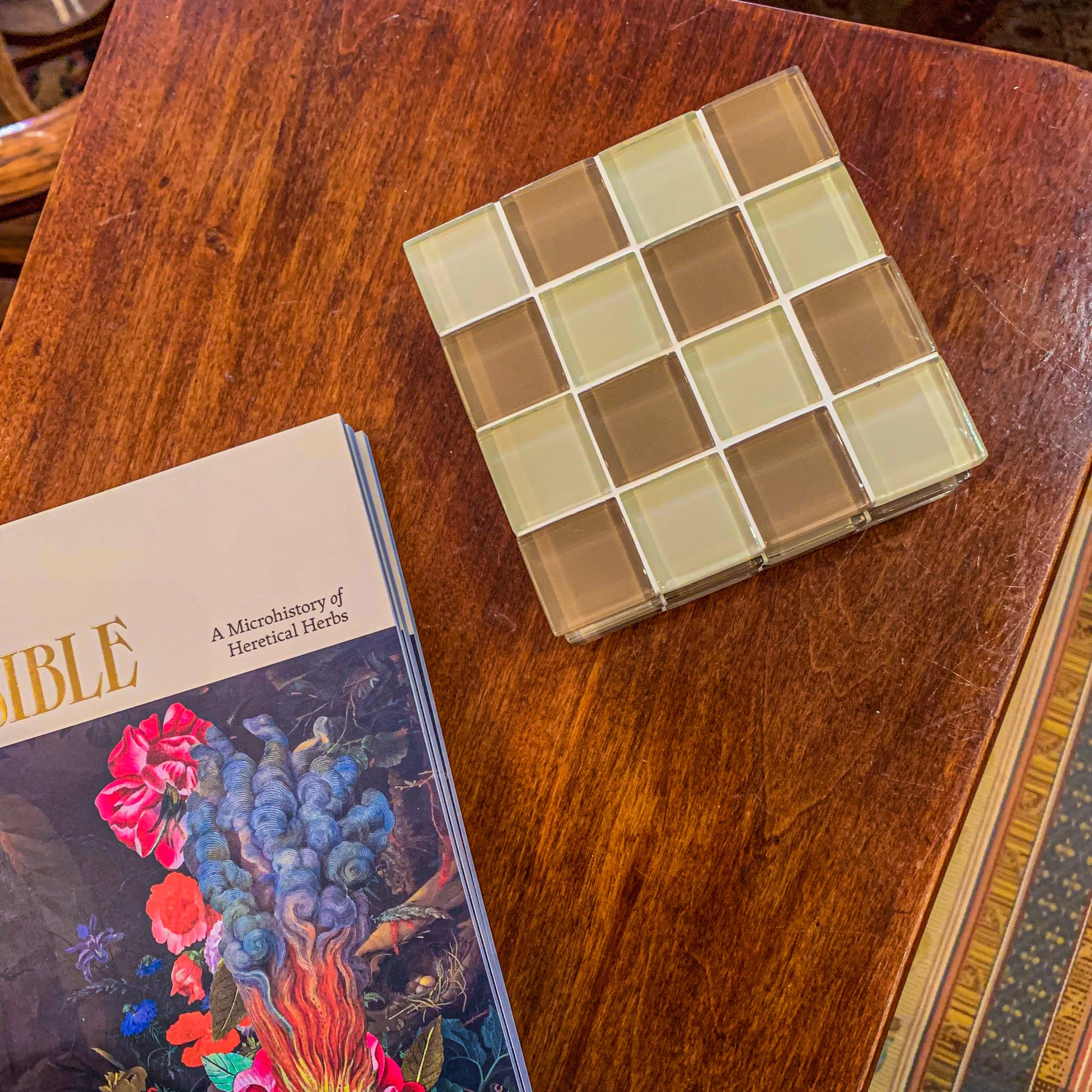 Subtle Art Studio Cafe Latte checkered coaster (tan & brown) on a wooden side table next to a book which reads “a microhistory of Heretical Herbs”