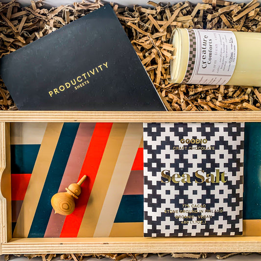 "Home Office" Gift box contains a masculine wooden tray with thick stripes and a red accent. There is a sea salt bar of chocolate,  a pack of Productivity Sheets, a wooden spinning top and a soy wax candle