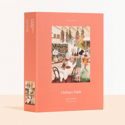 The Box of Ordinary Habit's "Home Flowering" 500 piece puzzle. Box is light pink and shows the puzzle picture which is a woman at her kitchen with flowers hanging from the ceiling and glass vases with dried flowers displayed around the room. Illustrated by Lida Ziruffo